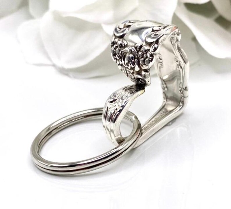 May include: A handcrafted silver key ring made from a repurposed spoon. The spoon's handle is bent to form a loop, and the spoon's bowl is decorated with floral patterns. A silver key ring is attached to the spoon.