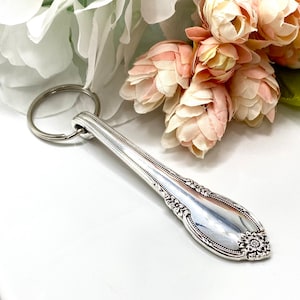 May include: Silverware keychain with a floral design on the handle. The keychain is made from a repurposed spoon and features a decorative floral pattern at the end. The spoon is attached to a silver key ring.