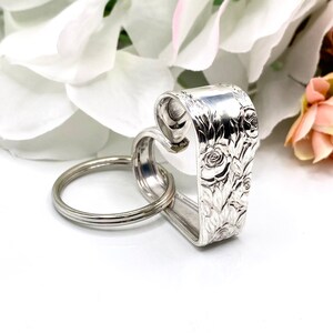 May include: A silver-toned metal keychain crafted from a bent spoon handle, featuring an embossed floral rose pattern on the exterior surface. The handle is shaped into a heart silhouette and is attached to a circular metal split ring for keys.