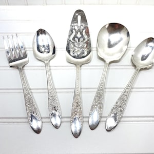 May include: A collection of vintage silver serving utensils, including a fork, spoons, and a pie server. The utensils have ornate floral detailing on the handles and are arranged against a white background.