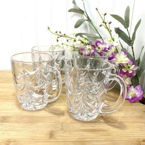 May include: A set of four clear glass mugs featuring a molded relief pattern of evergreen trees around the exterior. Each mug has a smooth, curved handle and a thick base, with the tree design extending from the bottom to the rim.