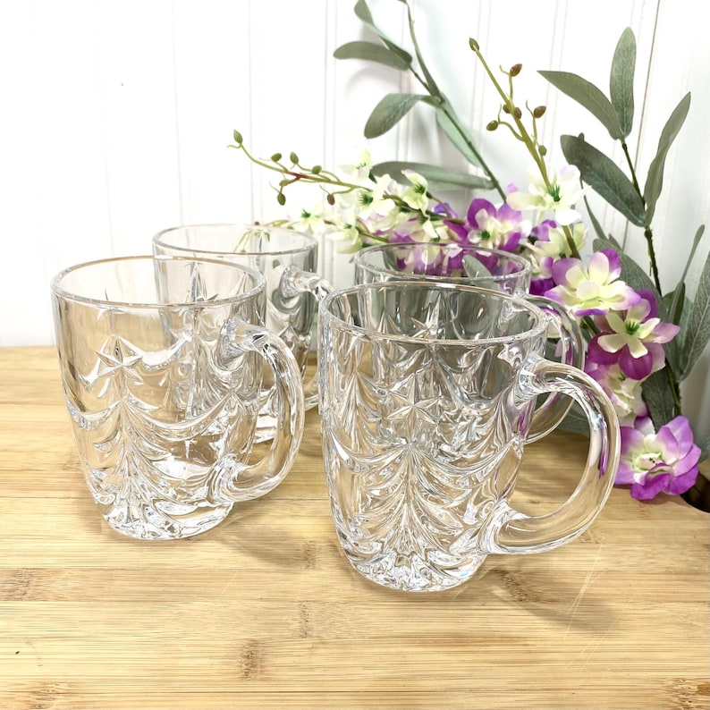 May include: A set of four clear glass mugs featuring a molded relief pattern of evergreen trees around the exterior. Each mug has a smooth, curved handle and a thick base, with the tree design extending from the bottom to the rim.