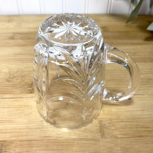 May include: A clear glass mug featuring a pressed leaf pattern on the exterior surface. The base of the mug displays a starburst design, and the handle is smooth and curved. The mug is positioned upside down, showcasing the intricate glass detailing.