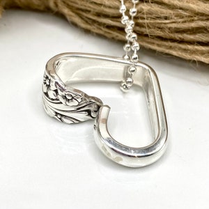 May include: A handcrafted silver pendant necklace. The pendant is made from a repurposed spoon, with a floral design on the spoon's bowl. The pendant is connected to a silver ball chain. The necklace is displayed on a white surface.