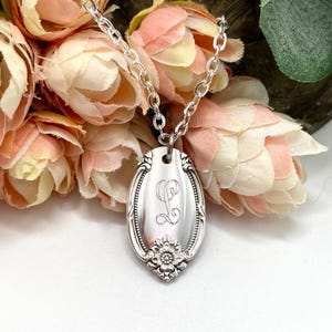 May include: A silver-colored spoon pendant necklace with an engraved letter "L" on an oval-shaped pendant. The pendant has a floral design at the bottom and is suspended from a delicate chain. The background features soft pink and peach flowers.