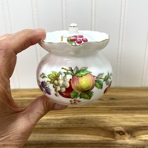 May include: A white porcelain sugar bowl with a scalloped rim and a matching lid. The body features a colorful botanical illustration of various fruits including apples, grapes, and blackberries, with a small cluster of cherries on the lid.