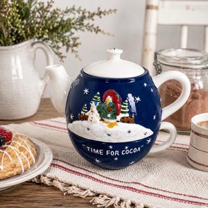 May include: A dark blue ceramic teapot with a white lid and handle, featuring a raised relief of a snowman in a green sweater and red hat. The text "TIME FOR COCOA" is printed in white on the front, surrounded by white snowflakes.