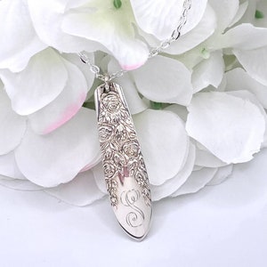 May include: A silver necklace with a pendant featuring a floral design and an engraved letter "S". The pendant is shaped like a long, narrow oval.