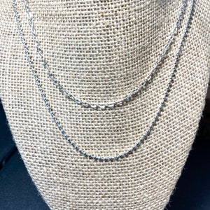 May include: Two silver necklaces displayed on a burlap-covered jewelry stand. The top necklace has a delicate chain link design, while the bottom necklace features small, evenly spaced beads. The necklaces are layered, creating a stylish and elegant look.