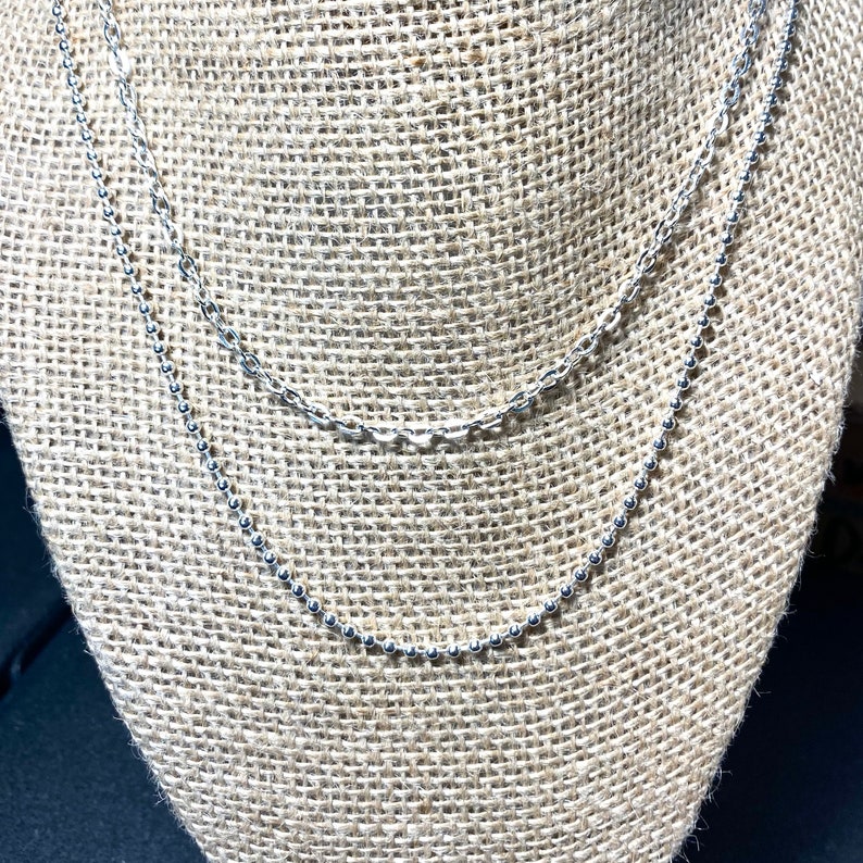 May include: Two silver necklaces displayed on a burlap-covered jewelry stand. The top necklace has a delicate chain link design, while the bottom necklace features small, evenly spaced beads. The necklaces are layered, creating a stylish and elegant look.