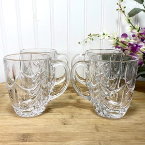 May include: A set of four clear glass mugs featuring a textured, embossed pattern of stylized evergreen trees and star shapes. Each mug has a sturdy handle and a smooth rim, crafted from transparent glass with a faceted, light-reflecting surface.