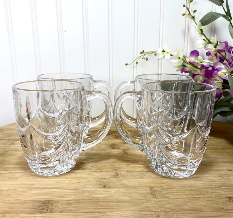 May include: A set of four clear glass mugs featuring a textured, embossed pattern of stylized evergreen trees and star shapes. Each mug has a sturdy handle and a smooth rim, crafted from transparent glass with a faceted, light-reflecting surface.