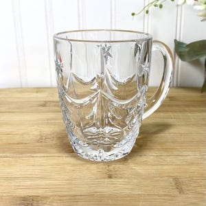 May include: A clear glass mug featuring a decorative pressed pattern of vertical arches and small star shapes. The mug has a smooth, rounded handle on the right side and a thick, sturdy base, showcasing a reflective, transparent finish throughout.