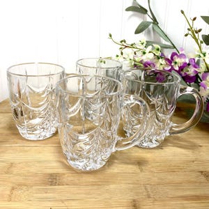 May include: A set of three clear glass mugs featuring a textured, embossed pattern of stylized Christmas trees and stars. Each mug has a smooth, curved handle and a cylindrical body, crafted from transparent glass with a reflective, faceted surface.