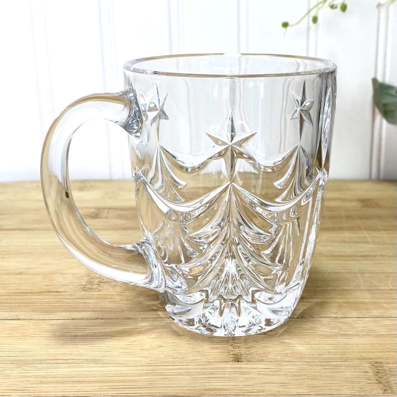 May include: A clear glass mug featuring a molded relief pattern of stylized evergreen trees and stars. The mug has a smooth, rounded handle and a wide, open top, with the intricate tree and star design embossed into the exterior surface of the glass.