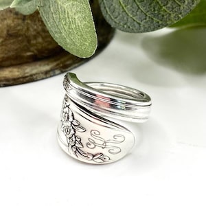 May include: A silver spoon ring with a floral design and engraved initials. The ring is made from a vintage spoon and is a unique piece of jewelry.