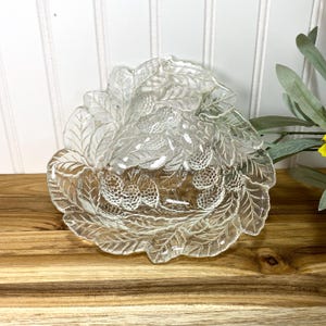 May include: A clear glass plate featuring a detailed embossed design of textured leaves and clusters of berries. The plate has a scalloped edge and a translucent finish, showcasing a botanical pattern that covers the entire surface of the dish.