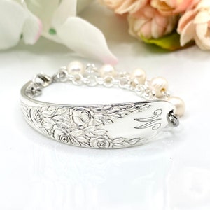 May include: A silver bracelet crafted from a spoon, featuring intricate floral detailing and a delicate chain of pearls. The spoon's handle is engraved with the letter "M". The bracelet is a unique piece of jewelry.