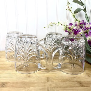 May include: A set of four clear glass mugs featuring a pressed starburst and leaf pattern. Each mug has a sturdy handle and a smooth rim. The glass is transparent with a reflective surface, showcasing the intricate embossed design on the exterior.