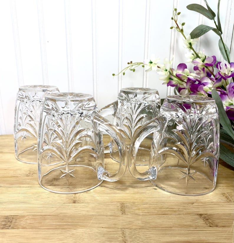 May include: A set of four clear glass mugs featuring a pressed starburst and leaf pattern. Each mug has a sturdy handle and a smooth rim. The glass is transparent with a reflective surface, showcasing the intricate embossed design on the exterior.