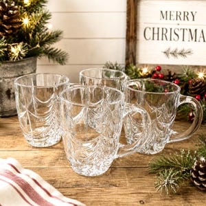 May include: A set of four clear glass mugs featuring a textured, embossed pattern of stylized evergreen trees around the exterior. Each mug has a sturdy handle and a smooth rim, designed for holding hot or cold beverages during the winter season.