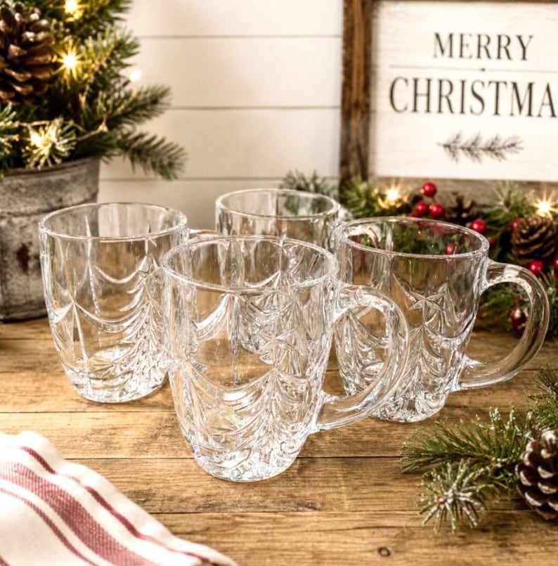 May include: A set of four clear glass mugs featuring a textured, embossed pattern of stylized evergreen trees around the exterior. Each mug has a sturdy handle and a smooth rim, designed for holding hot or cold beverages during the winter season.