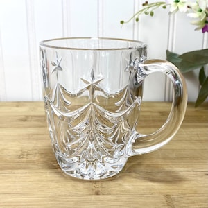May include: A clear glass mug featuring a molded relief pattern of stylized evergreen trees and stars. The transparent vessel has a smooth interior, a sturdy handle, and a textured exterior surface that catches the light, creating a decorative effect.