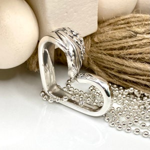 May include: A silver heart-shaped pendant necklace. The pendant is crafted from a spoon, with ornate floral detailing on the top curve. The necklace features a delicate ball chain. The background includes a white surface and a natural twine.