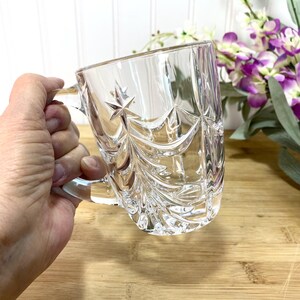 May include: A clear glass mug featuring a faceted, pressed pattern that resembles a stylized Christmas tree with a star at the top. The mug has a smooth, curved handle and a thick base, showcasing intricate geometric cuts that catch the light.