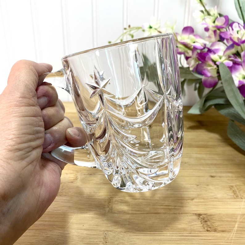 May include: A clear glass mug featuring a faceted, pressed pattern that resembles a stylized Christmas tree with a star at the top. The mug has a smooth, curved handle and a thick base, showcasing intricate geometric cuts that catch the light.