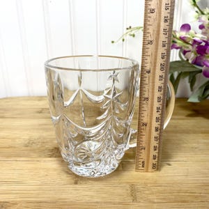 May include: A clear glass mug featuring a molded Christmas tree pattern with star accents. A wooden ruler is positioned vertically against the side of the mug, indicating a height of approximately 3.5 inches from the base to the rim.