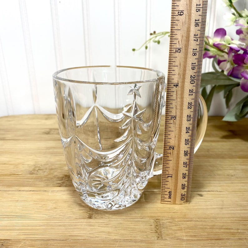May include: A clear glass mug featuring a molded Christmas tree pattern with star accents. A wooden ruler is positioned vertically against the side of the mug, indicating a height of approximately 3.5 inches from the base to the rim.