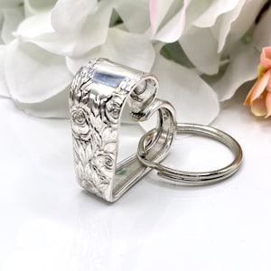 May include: A silver-toned metal keychain crafted from a bent spoon handle, featuring an intricate embossed floral rose pattern on the exterior surface. The handle is shaped into a heart silhouette and is attached to a circular metal split ring.