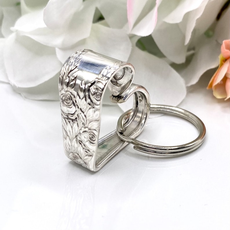 May include: A silver-toned metal keychain crafted from a bent spoon handle, featuring an intricate embossed floral rose pattern on the exterior surface. The handle is shaped into a heart silhouette and is attached to a circular metal split ring.