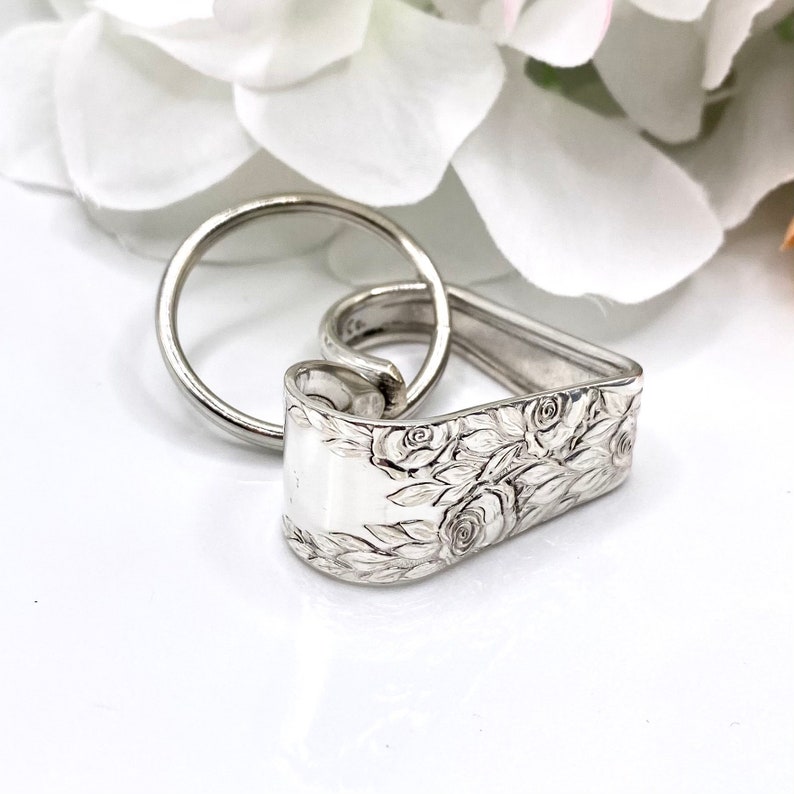 May include: A silver-toned metal keychain featuring a heart-shaped charm crafted from a bent piece of vintage silverware. The surface of the metal is embossed with a detailed floral pattern of roses and leaves, and a circular split ring is attached.