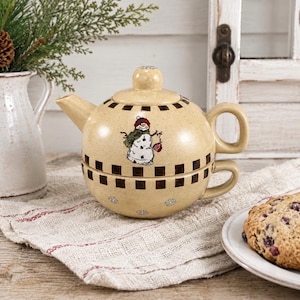 May include: A beige ceramic tea-for-one set featuring a teapot stacked on a teacup. The set has a speckled finish, a dark brown checkered border, and a central illustration of a snowman wearing a green scarf and holding a red ornament.