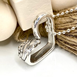 May include: A silver pendant necklace crafted from a repurposed spoon, featuring a floral design on the spoon's handle. The pendant is connected to a silver ball chain. The necklace is displayed on a neutral background.