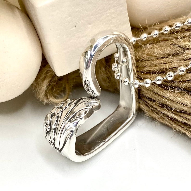 May include: A silver pendant necklace crafted from a repurposed spoon, featuring a floral design on the spoon's handle. The pendant is connected to a silver ball chain. The necklace is displayed on a neutral background.