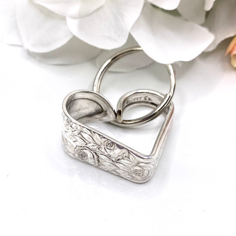 May include: A silver-toned metal keychain shaped like a heart, crafted from a bent spoon handle with an embossed floral pattern. The keychain includes a circular split ring, and the inner surface of the metal shows the text Silver Co. engraved.