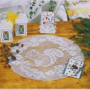 May include: A round white crocheted doily with a central squirrel motif, surrounded by two white metal lanterns with yellow ribbons, a small card featuring a robin, a small card featuring a squirrel, and several scattered brown pinecones.