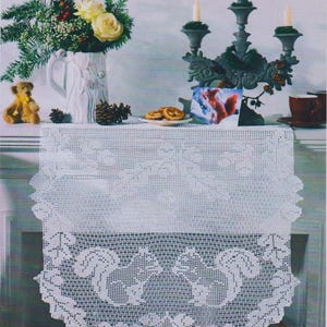 May include: White crocheted table runner with a delicate floral design. The runner is placed on a mantelpiece with a white pitcher filled with white roses and greenery, a candelabra with three lit candles, and a small plate of cookies.
