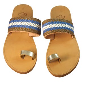 May include: A pair of brown leather sandals with a woven blue, white, and brown strap. The sandals have a silver toe ring and are made in Greece.
