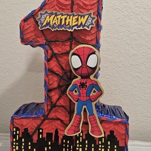 May include: A number one shaped piñata covered in red fringed paper with black web patterns. It features a cutout of a red and blue superhero character, a black city skyline silhouette with yellow windows, and the name Matthew in yellow text.