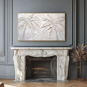 May include: A white textured painting of two palm trees with a gold frame. The painting is hanging above a white marble fireplace.