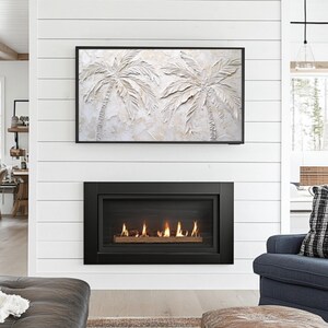May include: A black fireplace with a glass front and a realistic flame effect. The fireplace is set against a white shiplap wall with a framed artwork of palm trees above it.