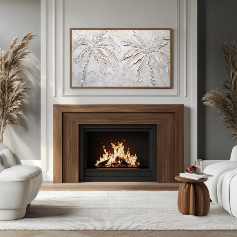 May include: A white textured wall art featuring two palm trees with a brown wooden frame. The art is hanging above a fireplace with a black surround and a roaring fire.