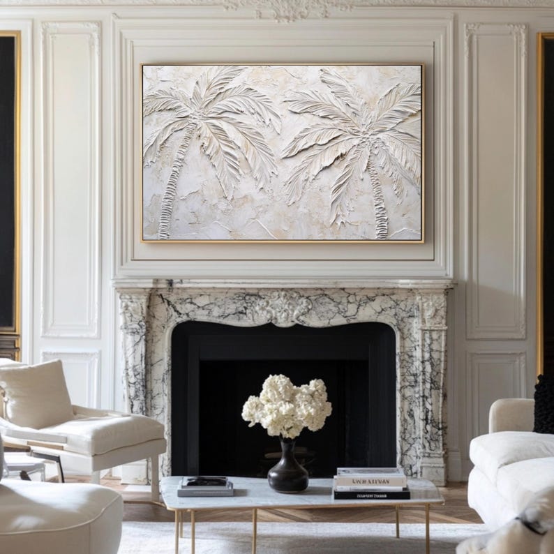 May include: A white textured painting of two palm trees with a gold frame. The painting is hanging above a marble fireplace in a white room.