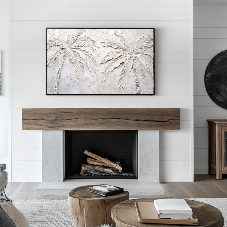 May include: A white textured painting of two palm trees with a black frame hanging above a fireplace with a wooden mantel and a black firebox. The fireplace is in a room with white shiplap walls and a wooden floor.