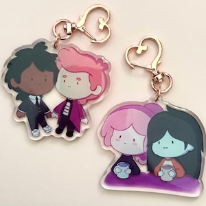 May include: A set of four cartoon character keychains. The first keychain features two characters, a boy with black hair and a boy with pink hair. The second keychain features two characters, a girl with pink hair and a girl with black hair. All characters are holding mugs.