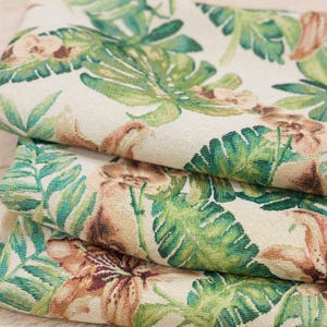 Op de afbeelding: Close-up van gevouwen stof met een tropisch bloemenpatroon. Het ontwerp heeft grote groene bladeren, bruine bloemen en kleinere groene bladeren op een crèmekleurige achtergrond. De stof lijkt geweven te zijn.